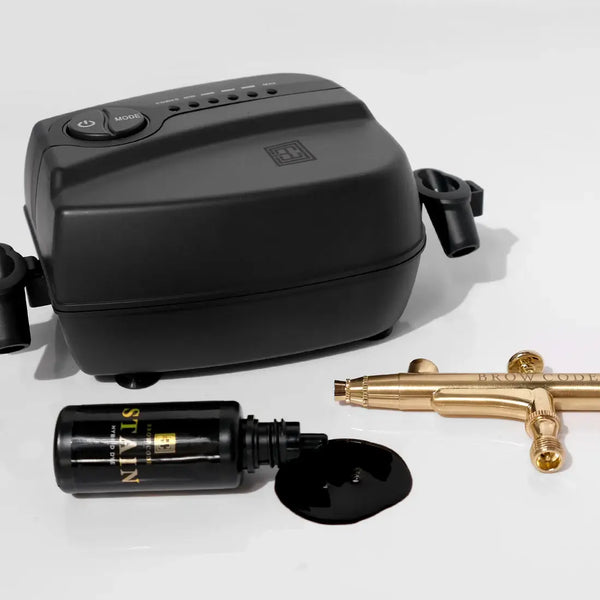 Brow Code Airbrush Machine with a black compressor, gold airbrush pen, and spilled black stain dye, showcasing its professional brow tinting capabilities