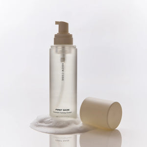 Brow Code First Base Ceramide Foaming Cleanser with the cap removed, standing in foamy lather on a reflective surface, showing the beige pump top