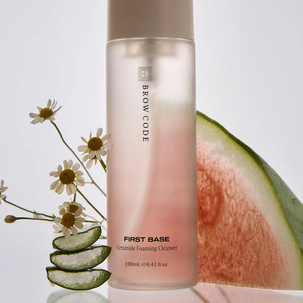 Brow Code First Base Ceramide Foaming Cleanser surrounded by natural ingredients, including aloe vera slices, chamomile flowers, and a fresh watermelon wedge