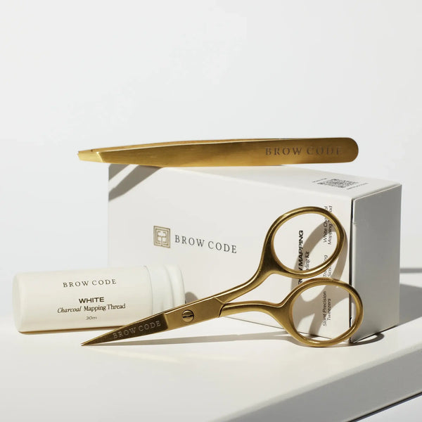 Close-up of Brow Code Mapping Kit with gold scissors, tweezers, and white charcoal thread resting on the branded packaging box