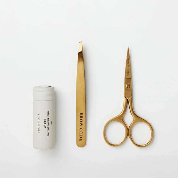 Flat lay of Brow Code Mapping Kit tools including white charcoal thread, gold slant tweezers, and gold trimming scissors on white