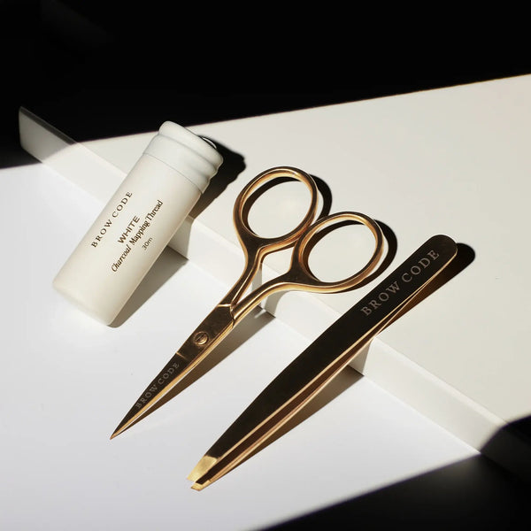 Brow Code Mapping Kit tools in shadowed light, featuring white charcoal thread, gold trimming scissors, and precision tweezers on display