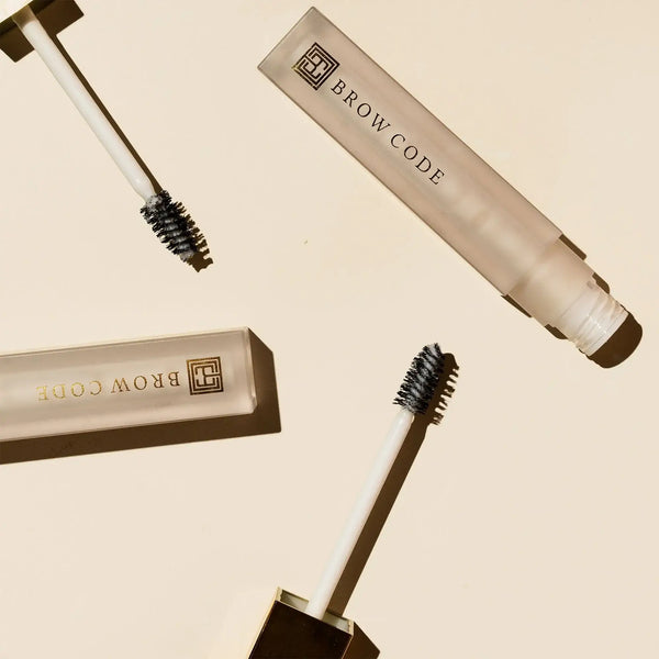 Flat lay of Brow Code Alias Brow Lamination Gel with frosted tubes and gold accents, featuring open applicators with product-coated spoolie brushes casting soft shadows