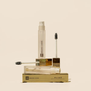 Brow Code Alias Brow Lamination Gel in a frosted tube with a gold cap, displayed with its applicator brushes and packaging, emphasizing its luxurious design
