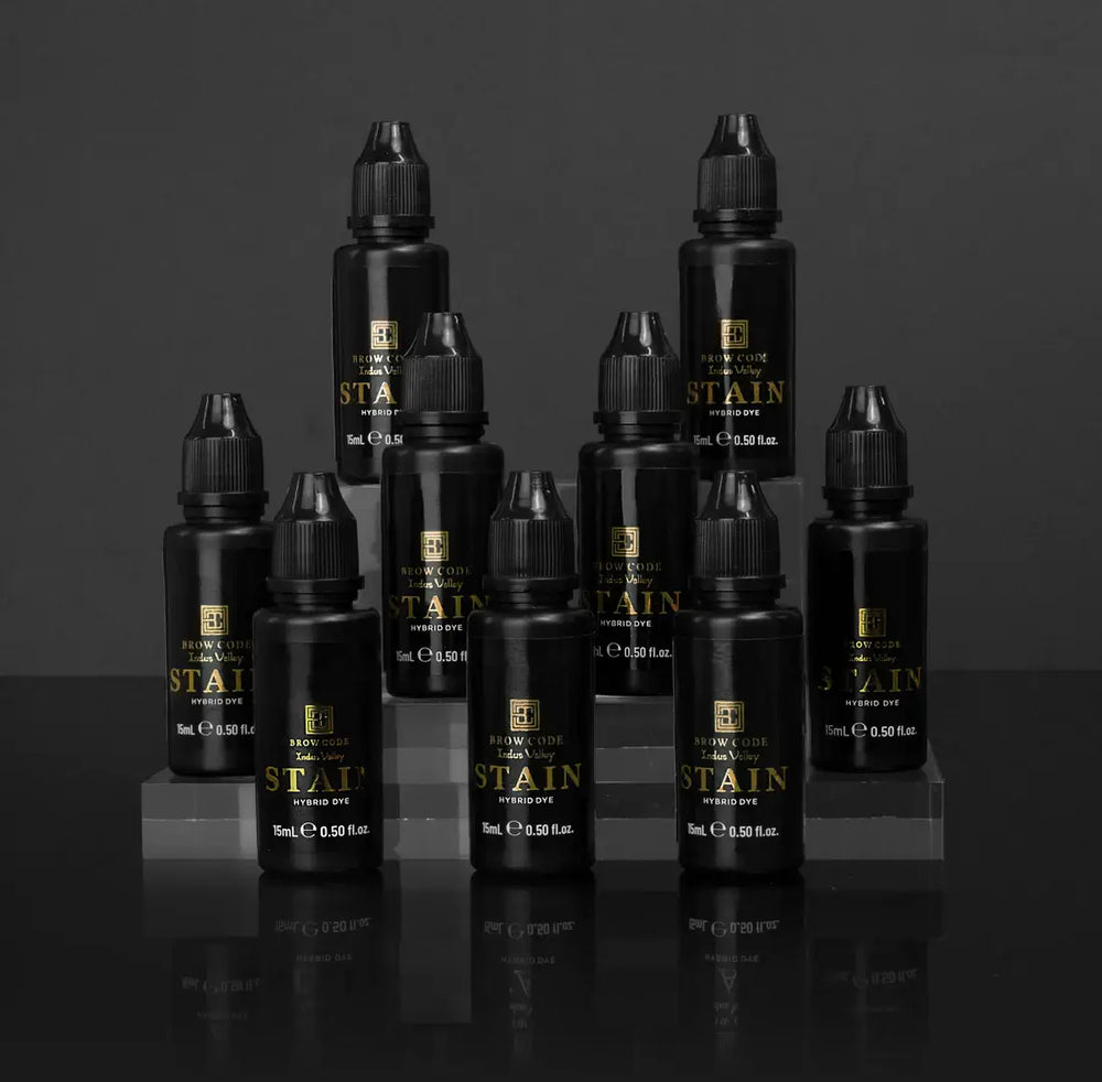 Brow Code Stain Hybrid Dye full collection displayed in a tiered arrangement, featuring 10 professional-grade 15ml bottles for brow tinting in multiple shades